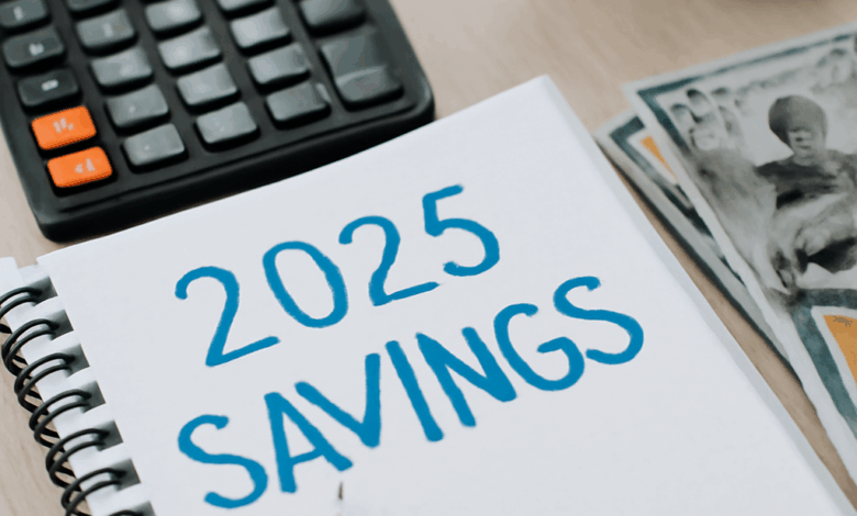 A close-up of a spiral-bound notebook with the bold heading '2025 SAVINGS,' along with a black calculator, financial documents with colorful bar graphs, scattered dollar bills, and a brass clock."